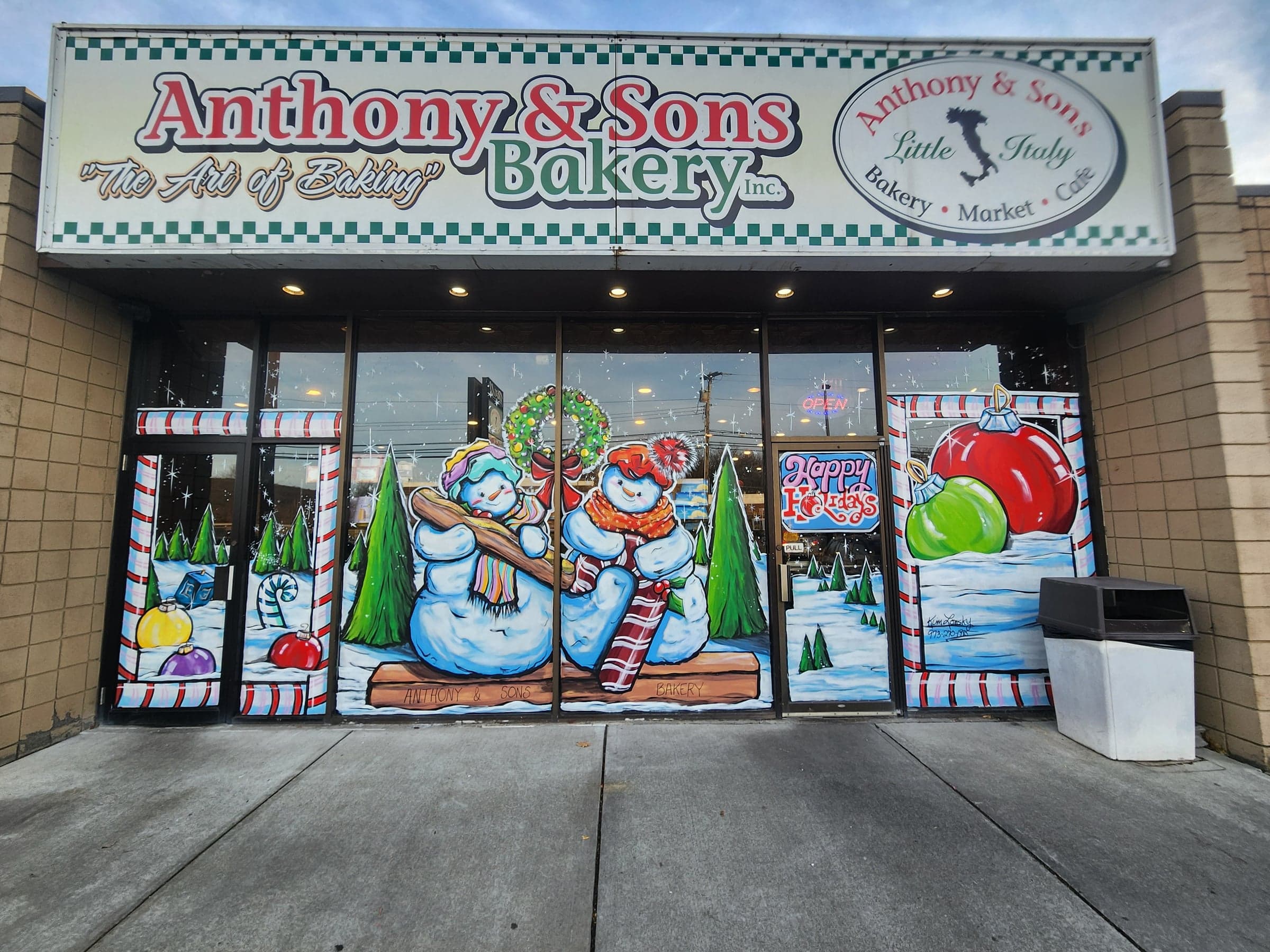 Hand-painted holiday storefront windows at Anthony & Sons Bakery — two snowmen on a bench among painted pines, a wreath, and giant ornaments, by Kim Lonsky